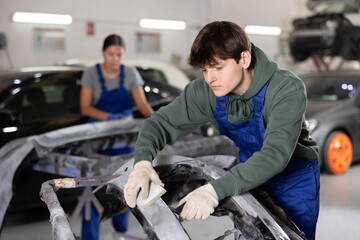 Young professional auto body technician in blue overalls and casual green hoodie, engaged in restoring damaged cars, expertly sanding removed bumper with sandpaper to smooth surface before painting