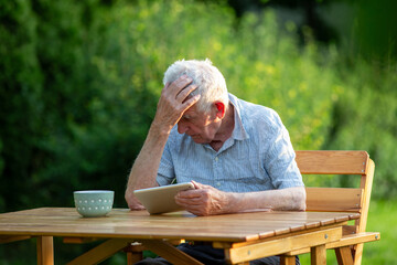 Old man holding head while looking away from tablet