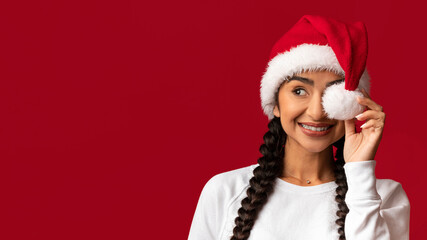 Obraz premium A joyful young woman smiles brightly while playfully holding a Santa hat pom-pom near her nose. The vibrant red background enhances her festive spirit and cheerful expression.