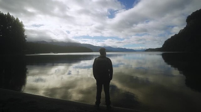 Rear view of a male hiker standing on a lakeshore looking at a peaceful mountain landscape with cloud reflections on the water.