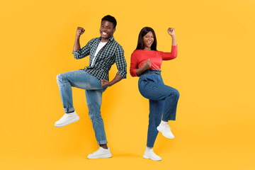 Fototapeta premium Two young adults express happiness by dancing together in playful poses. They wear casual clothes and enjoy the moment against a bright yellow background.