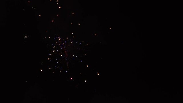 Colorful fireworks burst in the night sky, transitioning from bright sparks to vibrant blue and finally to a dazzling display of golden sparks illuminating the darkness