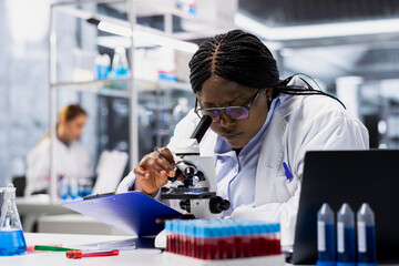 Female scientist in lab examining stained tissue sections on glass slide under microscope. African american woman checking cellular structure integrity for pathology report preparation