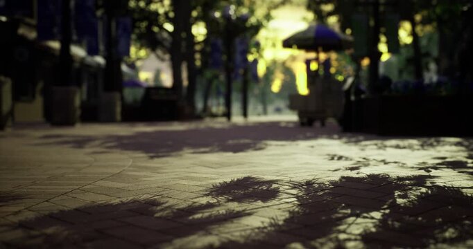 A quiet city park shows long shadows cast by trees as the sun sets, illuminating the path with warm hues. People stroll leisurely, enjoying the peaceful ambiance of the evening.