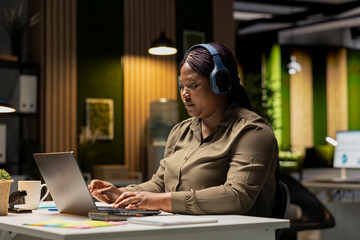 Female office worker managing daily tasks at night with focus music on headset, finding inspiration with ambience sounds to help with concentration and productivity. Woman multitasking.