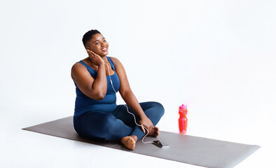 Fototapeta premium A woman sits on a yoga mat while enjoying music through earphones. She looks happy as she relaxes with a water bottle beside her. The setting is simple and bright.
