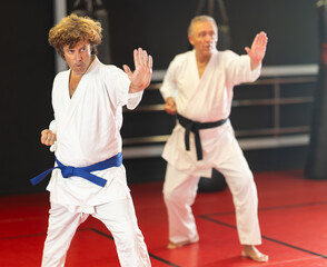 Focused middle-aged karateka in white kimono with blue belt performing kata with sensei, standing in attacking stance and practicing punch techniques in modern martial arts dojo..