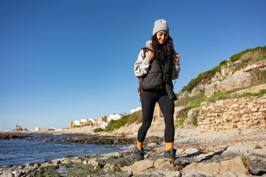 Young female hiker walking on rocky seashore with backpack, coastal travel lifestyle, outdoor adventure, freedom concept, active tourism, blue sky, seaside landscape