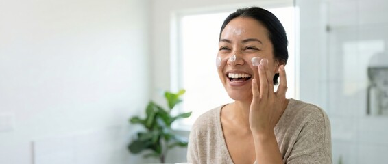 Asian woman applying face cream, laughing joyfully in bathroom Skincare routine, selfcare, and beauty concept