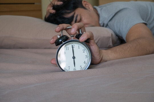 Tired young Asian man sleeping on bed and reaching hand to stop black alarm clock at 6 am. Sleepy male waking up early with headache or hangover in morning bedroom.