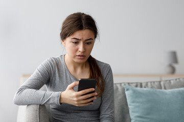 Worried sad shocked depressive attractive young european female looking at phone at home interior,...