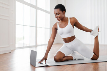 Fototapeta premium Athletic African woman doing fitness exercises on floor yoga mat, stretching feet and leg, following online sports tutorial at home. Lady using laptop to stay fit and healthy during covid 19 lockdown