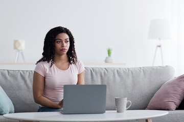 Problems in work and education at home during covid-19 quarantine, creating idea. Serious pensive young african american woman looking at empty space, in living room interior with laptop at workplace