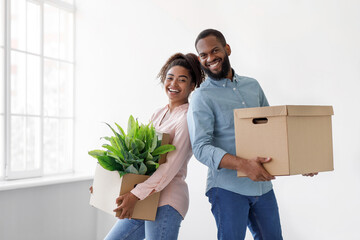 Smiling young african american husband and wife holding cardboard boxes with plant in pot, on white wall background. Sweet home, moving to new apartment, married couple is relocation in own flat