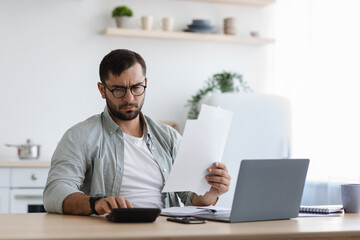 Serious busy sad adult european guy with beard in glasses counts taxes on calculator with laptop,...