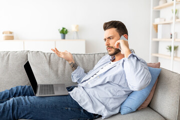 Emotional guy suffering problems with laptop, calling to customer support while sitting on couch in...