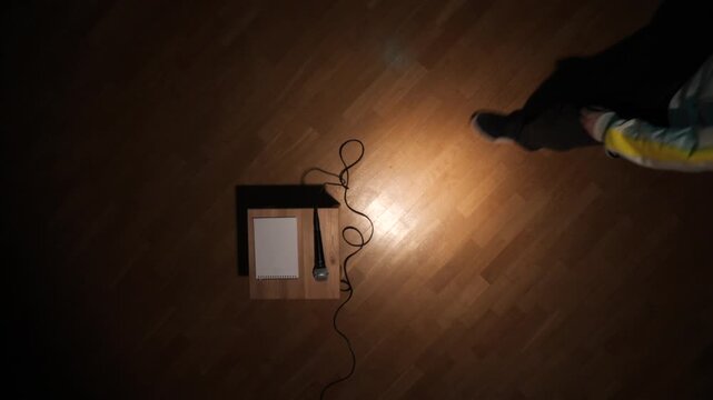 Spotlight illuminating a microphone and blank notebook on a small wooden stand, connected by a winding cable on a parquet floor, suggesting a stage for a performance