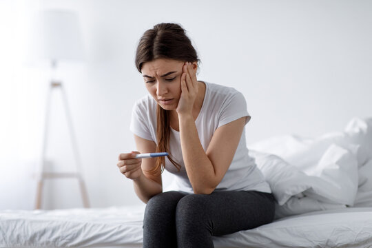 Depressed sad unhappy young pretty european woman looking at negative home test sitting on bed in bedroom interior alone, free space. Unwanted pregnancy, infertility, stress and health problems