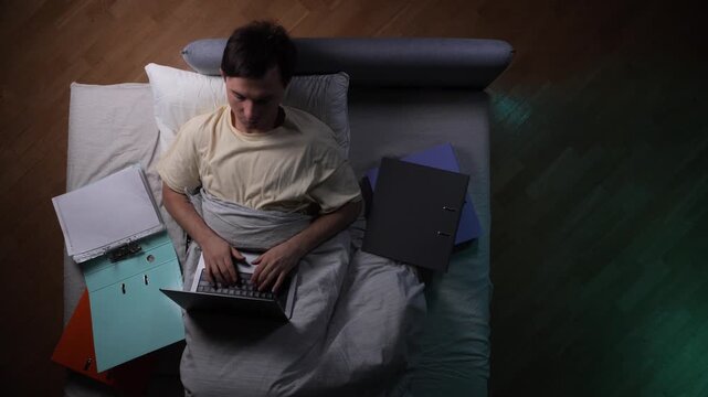 Young man lying in bed at night, typing on a laptop and surrounded by work files, representing burnout, overwork, and sleep deprivation due to demanding professional responsibilities
