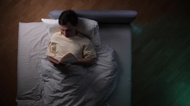 Man lying in bed under a duvet late at night, reading a book as he struggles with sleeplessness and an inability to fall asleep, a common sign of insomnia