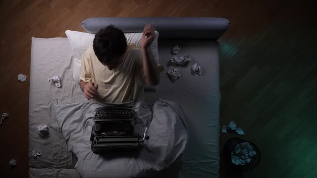High angle view of a man sitting in bed at night with a typewriter, surrounded by crumpled paper, symbolizing insomnia, frustration, and the struggle of creative inspiration