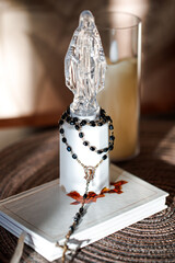 A clear statue of a religious figure stands on a table next to an open Bible. A rosary made of black beads is resting on the Bible, creating a simple but meaningful scene.