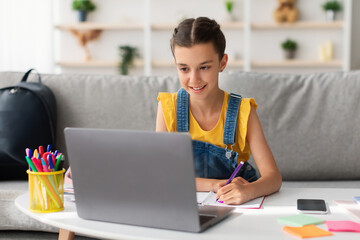 Portrait of smiling little girl drawing at home, using pc, happy schoolgirl looking at computer screen have fun in living room painting picture, studying online on quarantine, holding marker