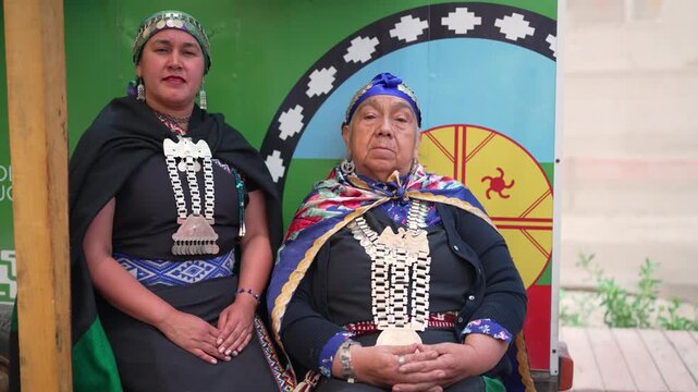 Two mapuche women wearing traditional jewelry and clothing are sitting and looking at the camera