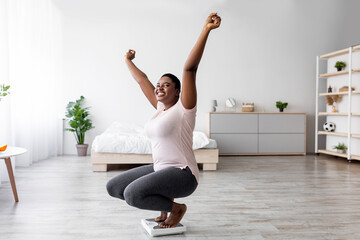 Successful weigt loss concept. Overjoyed plump black woman sitting on scales, raising hands up in...