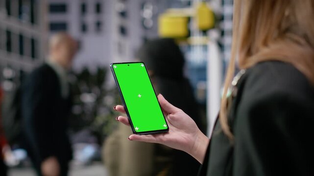 Vertical mockup screen on mobile phone held by a gen z young girl with backpack, diverse pedestrian group walking along the busy metropolitan streets during morning commute. Camera B.