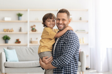 Fototapeta premium Happy family. Indoors portrait of positive caucasian man embracing his cute little son, holding him on hands, both smiling to camera at home interior