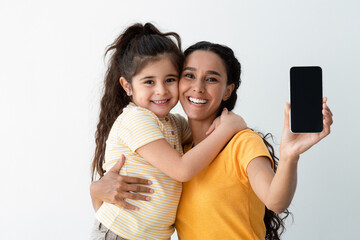 Cheerful Arab Mother And Little Daughter Demonstrating Smartphone With Black Blank Screen At...