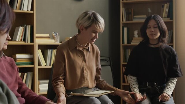Middle aged female psychotherapist mediating group therapy session with teenagers in counselling room then participants joining and raising hands celebrating unity and support