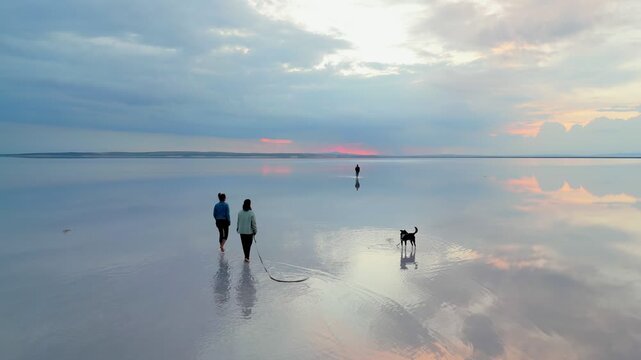 Friends walking barefoot with a dog on the salt flat reflecting the cloudy sky at sunset drone aerial view in Turkey