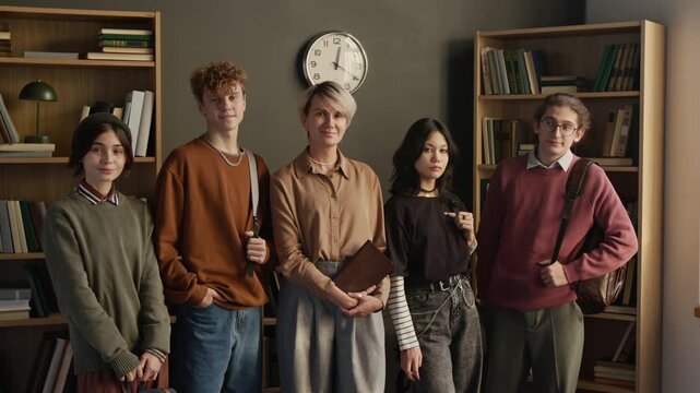 Portrait of adult female psychotherapist with notebook in hands smiling at camera while posing with five teenagers after group therapy session in counseling room, slow motion