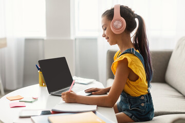 Side view profile portrait of girl in pink headset doing homework, sitting on couch at table using...