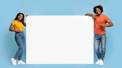 Stylish black people young man and woman pointing at empty advertising board between them and...
