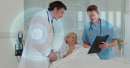 Examining doctor in lab coat leaning toward patient at bed, nurse in blue scrubs pointing clipboard