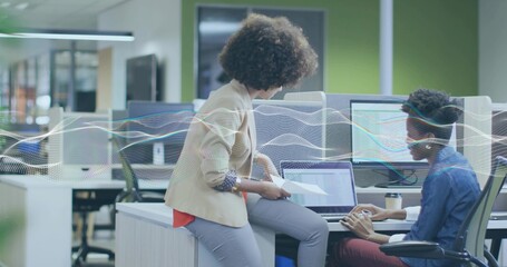 Collaborating two women in blazer and denim shirt reviewing laptop and printed pages at office