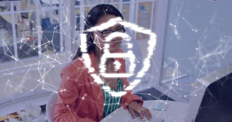 Typing Asian woman wearing coral blazer and glasses at office desk with keyboard and glowing symbol