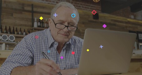 Writing senior man wearing checked shirt, reading glasses, using laptop and noting with pen at cafe
