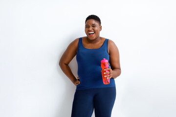 Naklejka premium A woman stands smiling while holding a water bottle. She is dressed in workout clothes and stands against a plain background. The atmosphere suggests a focus on fitness and hydration.