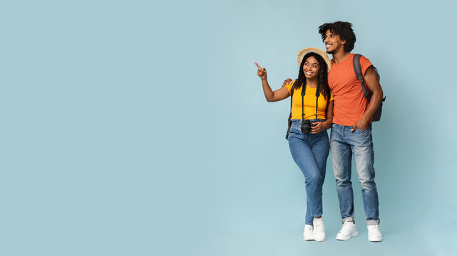 Happy millennial black couple tourists with backpacks and camera embracing and looking at copy space on blue studio background, choosing destination to travel, panorama. Travelling concept