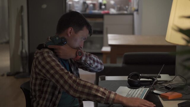 Cervical neck ache, nerve arthritis. Man studying late through night at laptop computer with cervical pain. Exhausted guy relaxing his neck using gun massager while overworking at notebook computer. 
