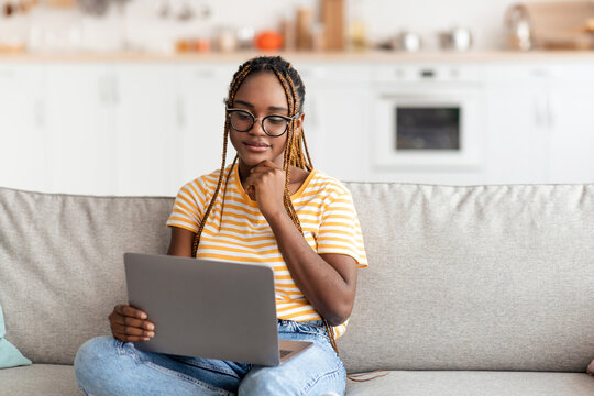 Attractive millennial african american woman with braids sitting on sofa, using modern notebook at home, unemployed young lady looking for job online, wearing eyeglasses, copy space