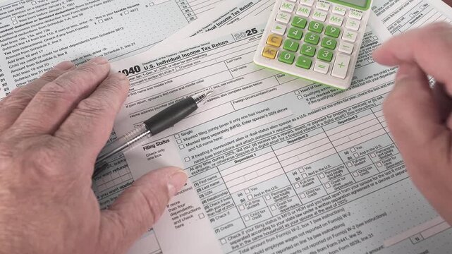 Hands are seen working on U.S. income tax 1040 forms while using a calculator. A pen is nearby for writing. The setting is a simple desk at home.