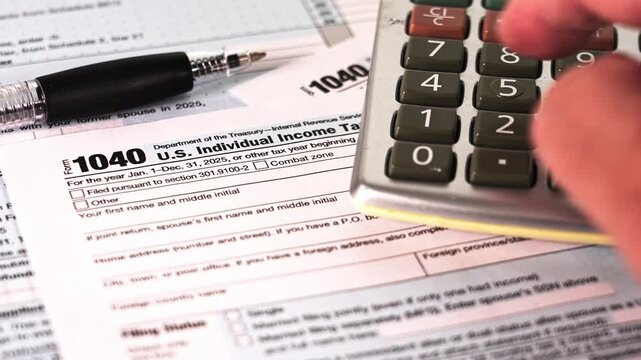 Person are seen working on U.S. income tax 1040 forms while using a calculator. A pen is nearby for writing. The setting is a simple desk at home.