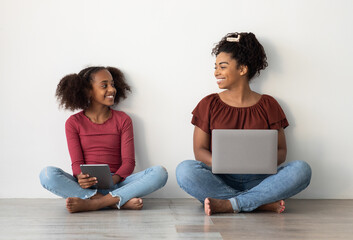 Happy black mom and teen kid using gadgets, sitting on floor over white wall, pretty african...
