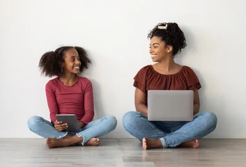 Fotobehang Diamanten Happy black mom and teen kid using gadgets, sitting on floor over white wall, pretty african american woman holding laptop and looking at her cute daughter with digital tablet, time together  © Prostock-studio