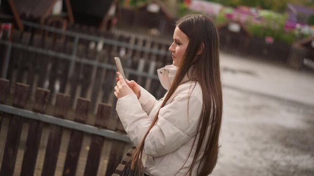 woman texting by wooden railing, overcast waterfront scene with raindrops on puffer jacket, long braided hair, blurred floral planters, reflective mood, urban riverwalk, closeup profile, scrolling